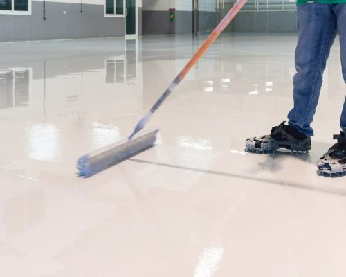 A contractor in gloves pours amber-colored epoxy resin from a metal bucket onto a garage floor, beginning the process of creating a durable, glossy protective coating.