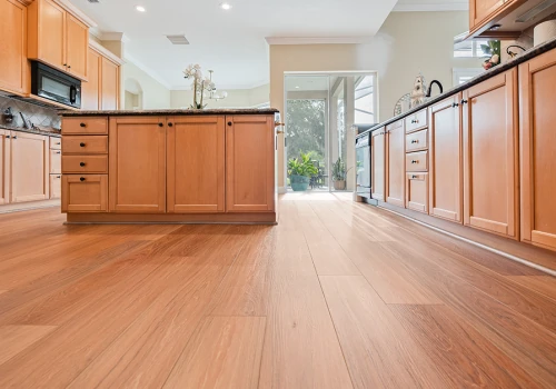 Rich, medium-brown wood-grain vinyl plank flooring complements natural oak cabinets and dark countertops in a bright, open kitchen with views to the outdoors.
