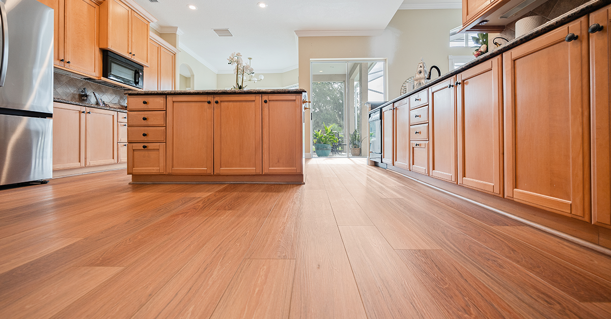 Rich, medium-brown wood-grain vinyl plank flooring complements natural oak cabinets and dark countertops in a bright, open kitchen with views to the outdoors.