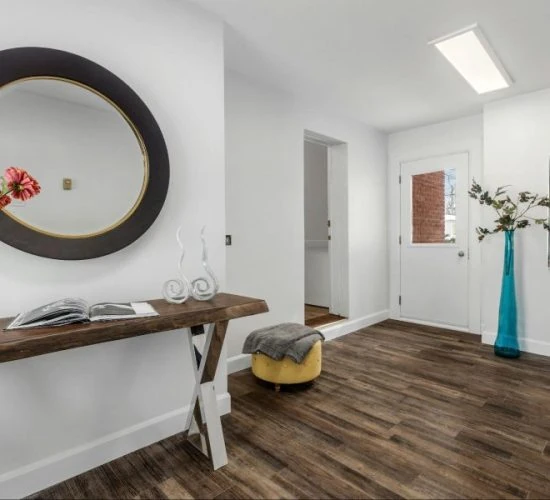 Rich, dark wood-grain laminate flooring anchors a contemporary entryway featuring a rustic wood console table, round mirror, yellow ottoman, and minimalist decor.