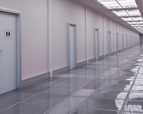 Sleek, large-format gray tile flooring extends down a clean, well-lit commercial hallway lined with white doors, one marked with a restroom symbol, under a skylight ceiling.