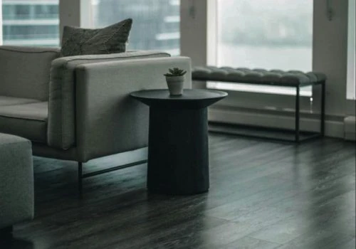 Sleek, dark wood-style flooring anchors a minimalist living space with gray sofas, black side tables, and large windows offering a city view — ideal for evaluating contractor quality.