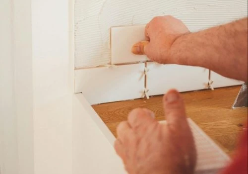 A skilled installer’s hands carefully place white rectangular tiles onto a wall with adhesive, using plastic spacers to ensure even grout lines — highlighting precision craftsmanship.