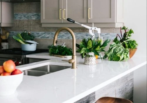 Sleek kitchen featuring a textured blue-grey mosaic tile backsplash behind a double sink, paired with gold fixtures, white countertops, and fresh produce for a stylish, functional look.