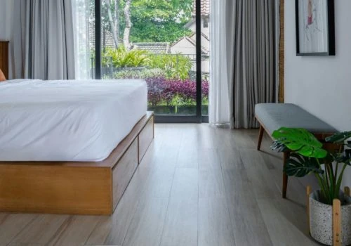 Calm bedroom featuring light gray wood-patterned vinyl flooring, a low wooden platform bed, and large glass doors opening to lush greenery — illustrating a finished, stress-free result.