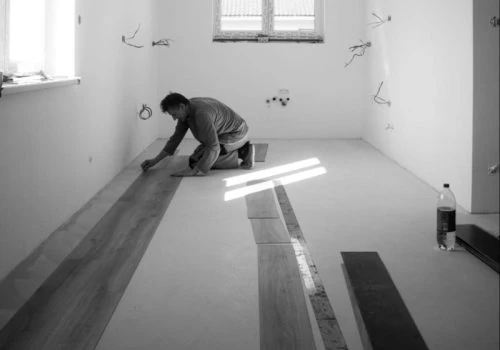 Monochrome image of a contractor kneeling to lay laminate flooring planks in a bright, unfurnished room with exposed electrical outlets and natural light from a window.