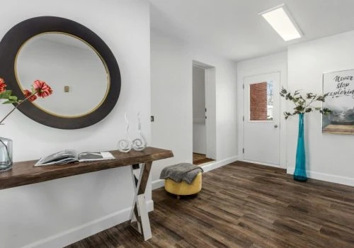 Rich, dark wood-grain laminate flooring anchors a contemporary entryway featuring a rustic wood console table, round mirror, yellow ottoman, and minimalist decor.