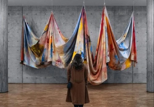 A visitor admires colorful fabric art installations in a minimalist gallery space featuring warm wood-style flooring, exposed concrete walls, and industrial columns.